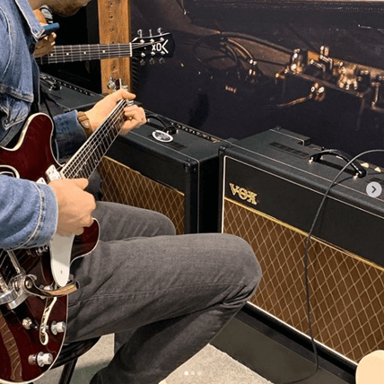partial view of two men playing electric guitars in front of VOX amplifiers