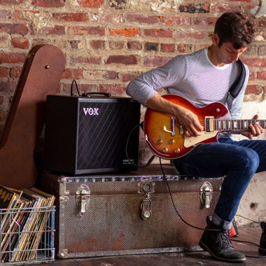 Vox Cambridge50 guitar modeling amp sitting on a trunk to the left of a man playing an electric guitar