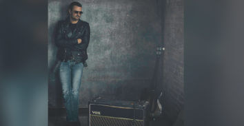 man leaning on wall behind VOX amplifier and beside electric guitar