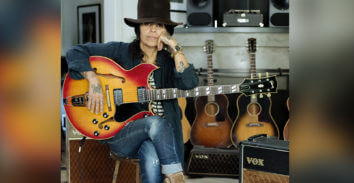 seated female musician holding electric guitar in front of VOX amplifiers and multiple acoustic guitars