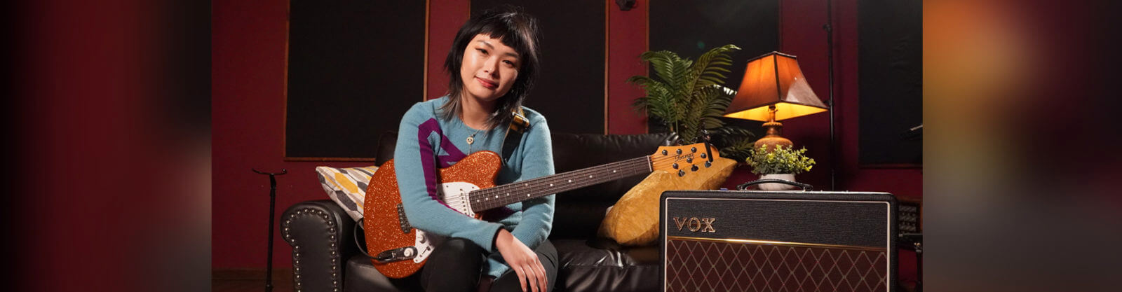 woman with electric guitar seated on leather sofa beside VOX amplifier
