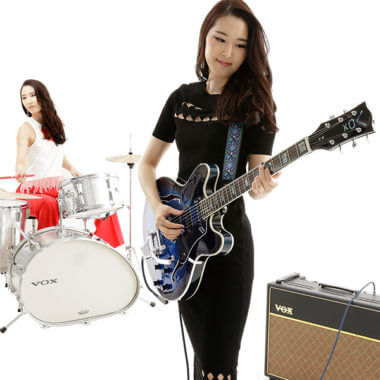 woman holding electric guitar with woman playing drums in background