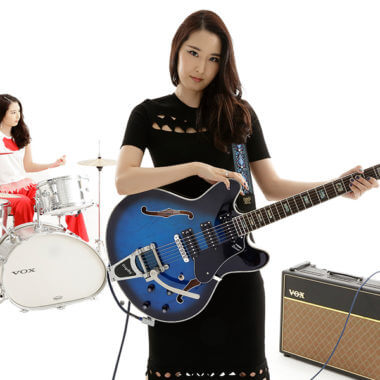 woman holding electric guitar with woman playing drums in background