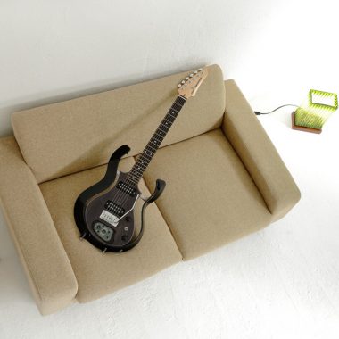 black VOX electric guitar on couch between small table and lamp